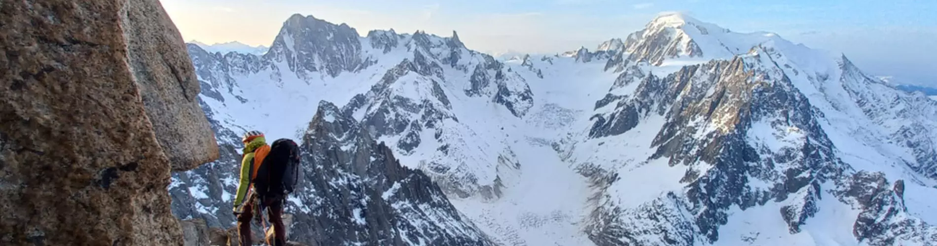 Een bergbeklimmer met een helm en uitrusting staat op een rotsrichel hoog boven besneeuwde bergen, vastgemaakt aan een veiligheidstouw, met op de achtergrond een panoramisch uitzicht op met sneeuw bed